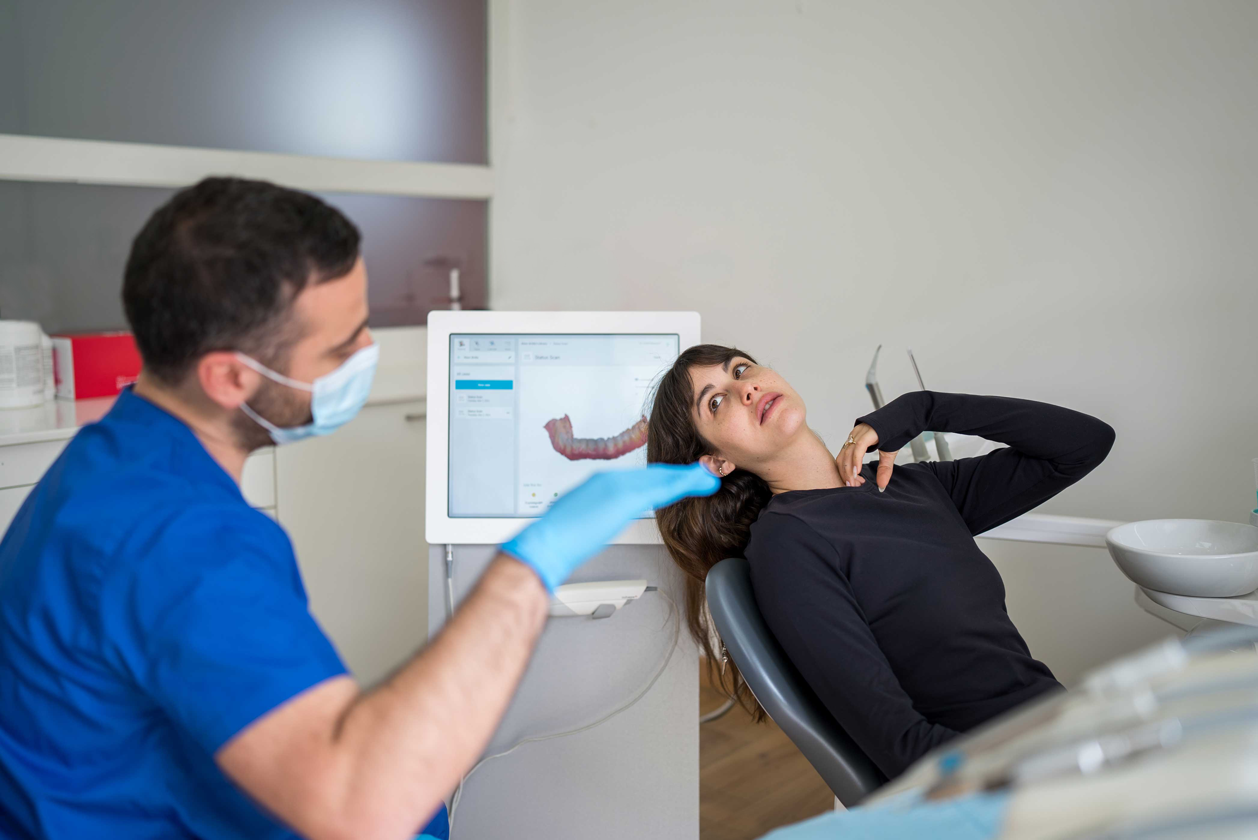 طبيب أسنان يجري مسحاً لأسنان المريض في عيادة متخصصة ويظهر شكل الأسنان على شاشة صغيرة في الخلف